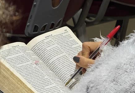 Bíblia aberta sobre o colo e uma mão anotando com caneta durante a aula